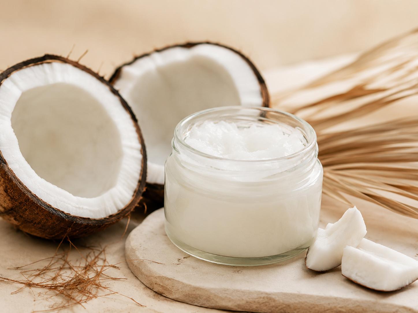 Clear glass jar of coconut oil beside fresh coconut halves and coconut pieces for dry scalp care